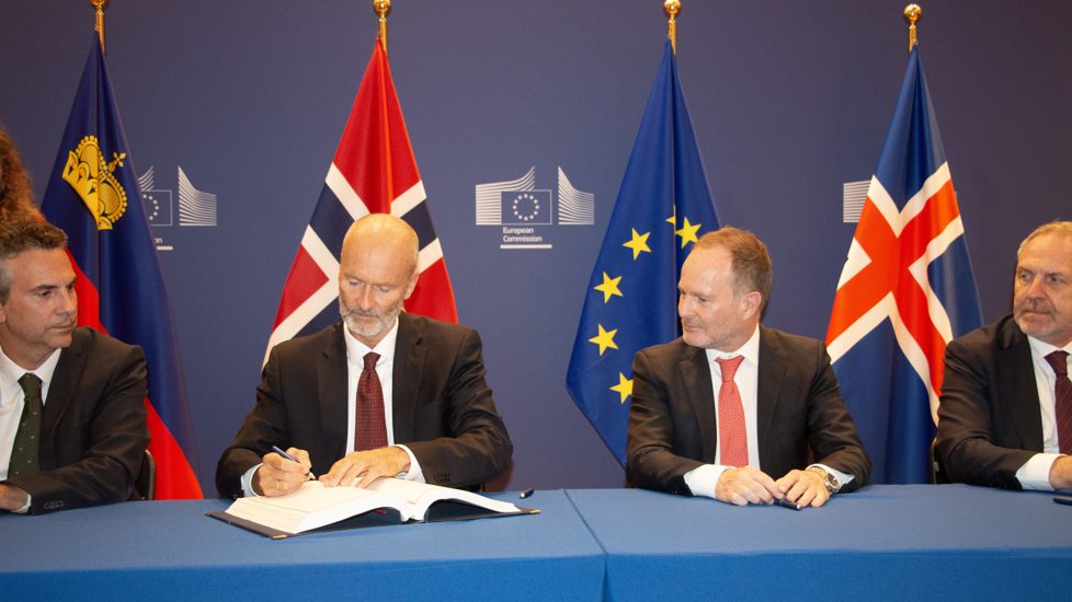 Bilde av fire ambassadører sittende for å signere avtale med flagg i bakgrunnen