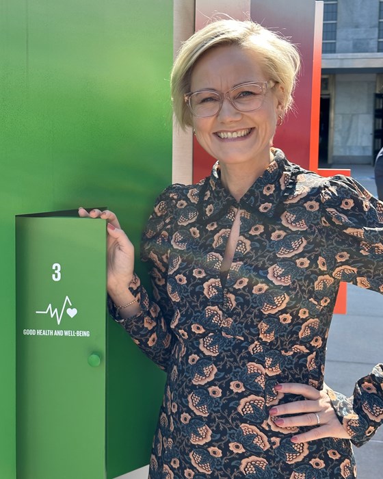 Bilde av helseminister Kjerkol med grønnbakgrunn og en åpen luke med bærekraftsmålet om god helse skrevet på.