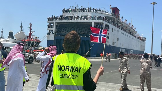 Bilde av ambassadeansatt i refleksvest med norsk flagg som ser på ferge med flere personer på dekk