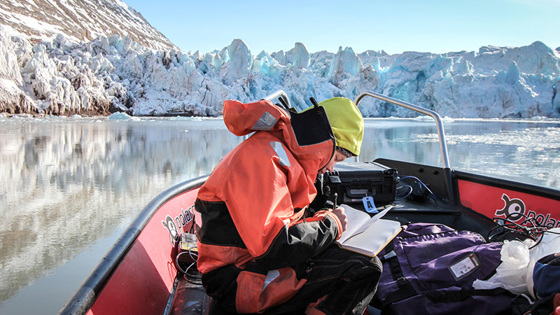 Bilde av person i oransj kjeledress på båt med isfjell rundt