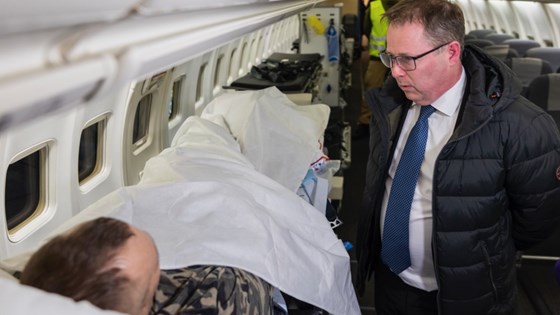 Bilde av forsvarsministeren som prater med pasient som ligger i seng på flyet